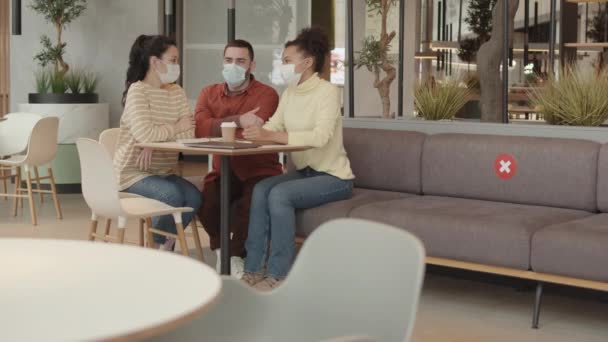 年轻多样的多民族同事戴着医疗面罩 坐在现代餐馆的餐桌旁 谈论大流行病期间的会面 — 图库视频影像