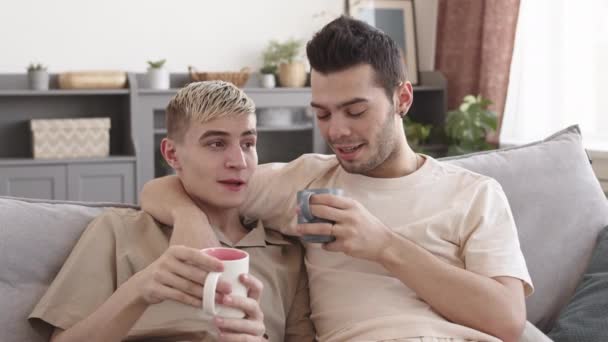 中等比例的年轻的金发白人男子和他的男朋友穿着休闲装坐在客厅的沙发上 边喝咖啡边聊天 — 图库视频影像