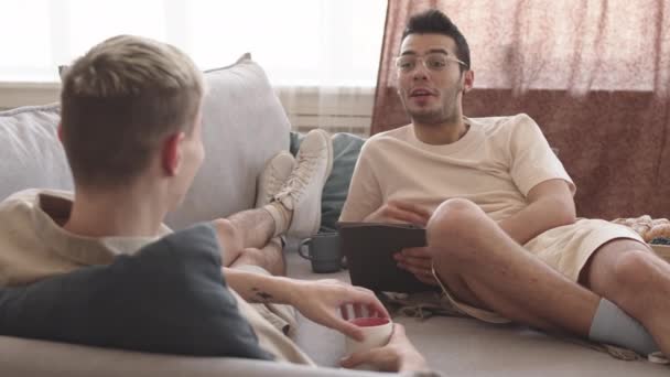 年轻的黑头发白人男子和他的男朋友一起躺在沙发上 用数码平板电脑从网上给他读有趣的信息的背景图 — 图库视频影像
