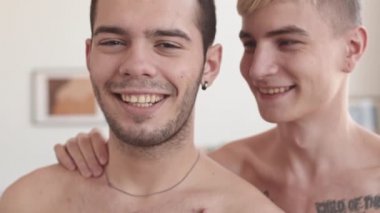 Siyah saçlı, çıplak gövdeli genç bir adamın yakın portresi dairede dikilip kameraya bakarken sevgilisi ona sarılıyor.