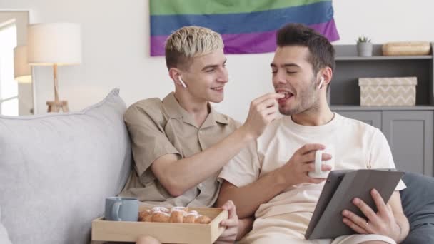 两个白人年轻人坐在家里的沙发上 用无线耳机和平板电脑看电影 金发男人用蓝莓喂他的男朋友 — 图库视频影像