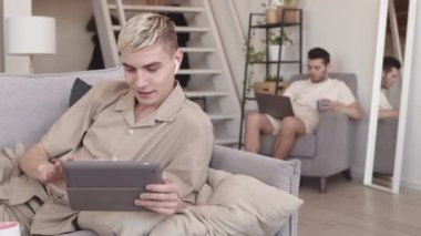 Erkek arkadaşı dizüstü bilgisayarla arka planda çalışırken orta boy sarışın beyaz adamın dijital tablette video izleyip kablosuz kulaklık kullandığı bir fotoğraf.