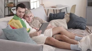 Evdeki kanepede sarılan iki beyaz genci kilit altına almak. Koyu renk saçlı adam kulaklıklarını çıkarıp dijital tablette video izlerken erkek arkadaşına takıyor.