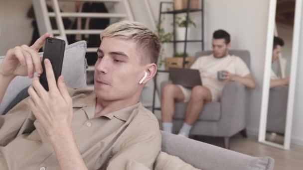 집에서 소파에 친구가 노트북으로 일하는 스마트폰으로 비디오 통화를 청년의 락다운 — 비디오