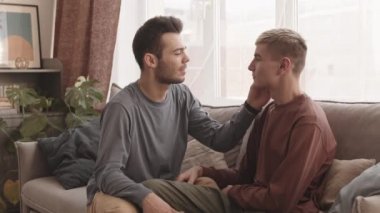 Orta boy, siyah saçlı, beyaz bir erkek ve sevgili erkek arkadaşının el ele tutuşup birbirlerine sevgiyle baktıkları bir fotoğraf.