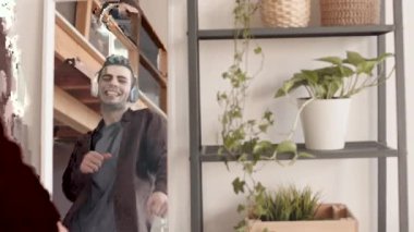 Genç, yakışıklı, koyu renk saçlı, günlük kıyafetler ve kulaklık takan, ayna ve evin önünde zımparalanan, müzik dinleyen ve dans eden bir adam.