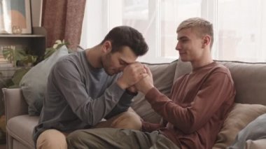 Siyah saçlı, günlük giysiler giyen, kanepeye oturmuş ve erkek arkadaşının elini tutan beyaz bir adam.