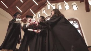 Mezuniyet elbisesi ve kepleri giyen, kapalı mekanda duran, gülümseyen ve parşömenlerle şakalaşan beş neşeli öğrencinin orta açısı.