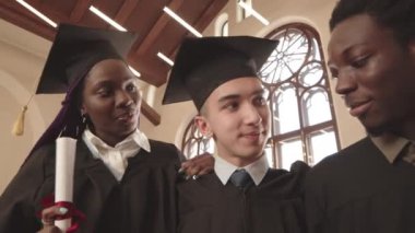 Erkek ve kadın Afrikalı, melez ve beyaz öğrencilerin göğüs hizasında mezuniyet elbisesi ve kepleri giyip, tomarları tutarak sırada bekleyip, birbirlerine sarılıyorlar.