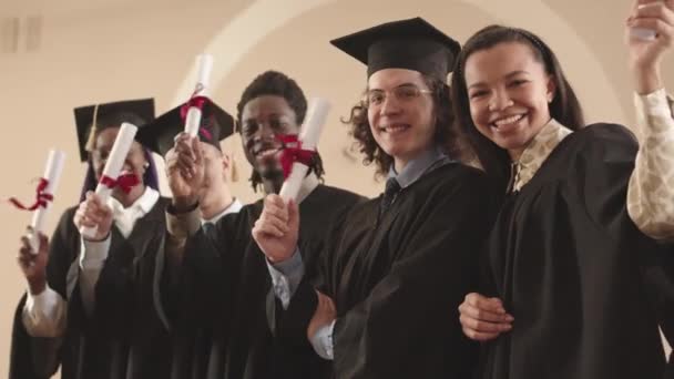 5名穿着毕业礼服 戴着毕业帽 站在室内 挥动卷轴 看着相机的年轻 快乐的学生的低角度中等生活质量 — 图库视频影像