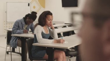 Orta uzunluktaki çoklu etnik çeşitlilikte öğrencilerin sınıflarda masalarda oturması, sınava yazı yazması, sonra da kameraların gözlüklü tanınmayan bir öğretmene odaklanması, onları izlemesi.