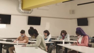 Orta uzunlukta, renkli maske takan, sınıftaki masalarda oturan, önemli sınavlara odaklanmış beş farklı etnik öğrenci.