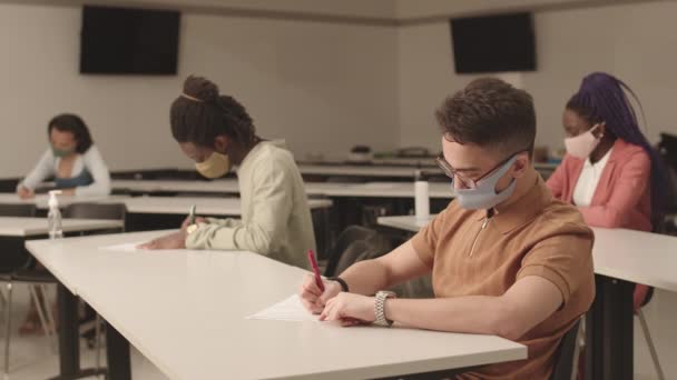 中等长度的男混血大学生戴着蓝色布面罩 参加了重要的考试 坐在教室的桌子前 面对着各种模糊的同学 — 图库视频影像