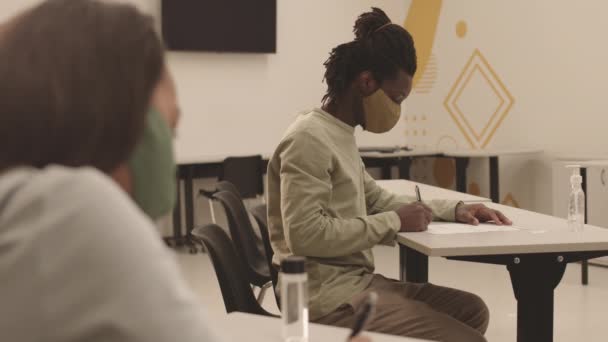 非洲男生戴着布面罩 坐在课桌前 参加考试 用纸写东西的中长侧视图 — 图库视频影像