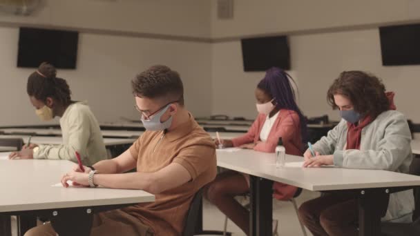 中长型多民族学生戴着定做的五颜六色的口罩 坐在课桌前 专心致志于重要的考试写作 — 图库视频影像