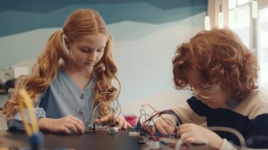 Orta boy iki kızıl saçlı, günlük kıyafetler giyen öğrencinin sınıfta masasında oturup robot üretirken çekilmiş bir fotoğrafı.