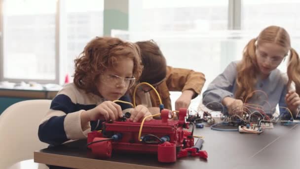 在机器人技术课上 3名白人学生一起坐在教室的课桌前 用不同的细节和电线制造机器人 — 图库视频影像