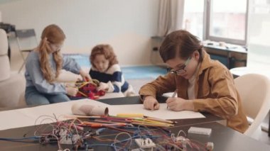 Sınıftaki iki sınıf arkadaşı arka planda robot çizerken beyaz öğrencilerin masaüstünde oturması ve resim çizmesi.