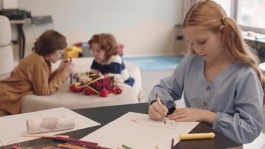 İki erkek sınıf arkadaşı arka planda robot yaparken, sınıfın masasında oturan ve resim çizen kızıl saçlı beyaz kız öğrencileri.