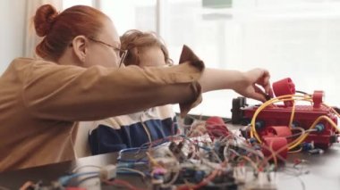 Genç, kızıl saçlı, günlük kıyafetler giyen, gözlüklü ve kıvırcık saçlı genç bir çocuğun birlikte oturduğu ve elektronik robotlar yaptığı bir yer.