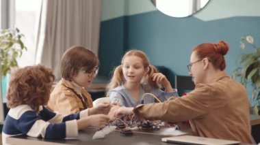 Orta boy beyaz kadın öğretmen, iki liseli kız ve erkek öğrenci masaüstünde oturmuş robot teknolojisi dersi sırasında elektronik robot yapıyorlar.