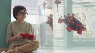 Sınıftaki su kabının başında oturan beyaz öğrencinin kilitli portresi. Joystick kullanarak suda yüzen robo botu kontrol ediyor ve sonra kameraya bakıyor.