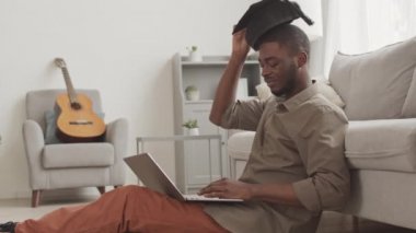 Mezuniyet şapkası takan genç bir Afro-Amerikalı dizüstü bilgisayarla evde oturuyor, online mezuniyet törenini bitiriyor, şapkasını çıkarıyor ve klavyede daktilo yazıyor.