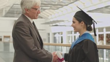 Yaşlı erkek üniversite hocası el sıkışıyor ve üniversite mezuniyet elbisesi ve şapkası giyen ve diplomasını elinde tutan melez bir kız öğrenciye sarılıyor.