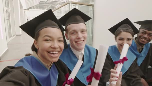 Střední záběr čtyř různých spolužáků, kteří na sobě mají vysokoškolské šaty a čepice, sedí spolu, dívají se na kameru a mávají diplomy, zatímco dělají selfie