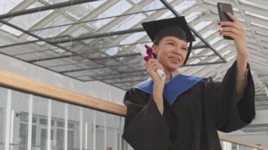 Orta boy, üniversite mezuniyet elbisesi ve şapkası giymiş genç, melez bir kız öğrencinin üniversite koridorunda durup telefonda görüntülü konuşma yaparken diplomasıyla el sallaması.