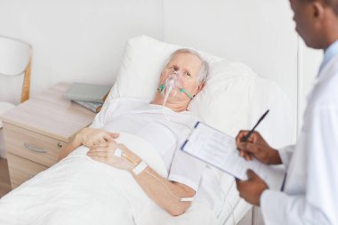 Hastane yatağında oksijen takviyeli maskeyle yatan ve Afro-Amerikan doktorlara bakan üst düzey bir adamın portresi.