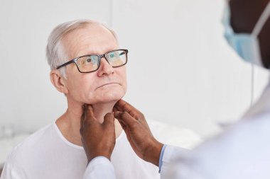 Tanımlanamayan Afro-Amerikan doktor hastane odasındaki muayene sırasında yaşlı bir adamın boynunu okşuyor.