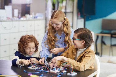 Portrait of three smiling children experimenting with electric circuits while building robots in engineering class at school