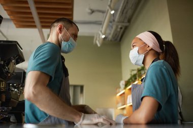 Side view portrait of two young workers wearing masks while standing at bar counter in cafe or coffee shop