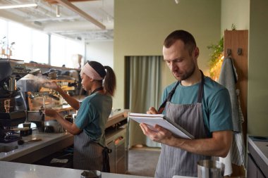 Waist up portrait of male barista wearing apron while working in coffee shop and holding clipboard, copy space
