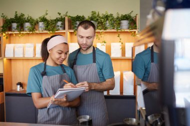 Waist up portrait of two young waiters wearing aprons and holding clipboard while doing inventory in cafe or coffee shop, copy space