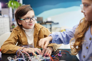 Portrait of boy and girl building robots and experimenting with electric circuits during engineering class at school, copy space