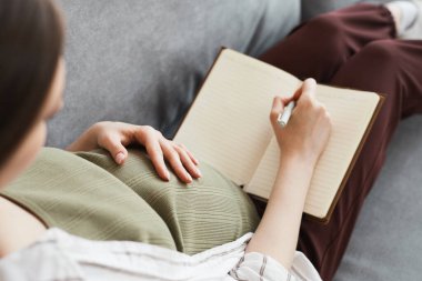 Rear view of pregnant woman lying on the sofa and making notes in her notebook she writing plans during her leisure time