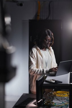 Karanlık sunucu odasında çalışırken dizüstü bilgisayar kullanan Afro-Amerikalı kadın ağ mühendisinin dikey portresi