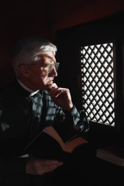 Serious senior priest sitting with Bible in confessional in the church clipart