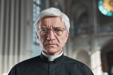 Kilisede dikilirken kameraya bakan gözlüklü kıdemli rahibin yakın çekimi.