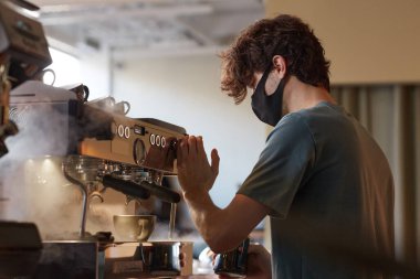 Side view portrait of young barista wearing mask while making fresh coffee in cafe or coffee shop, copy space