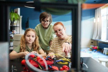 Front view portrait of female teacher pointing at 3D printer during engineering and robotics class at modern school, copy space