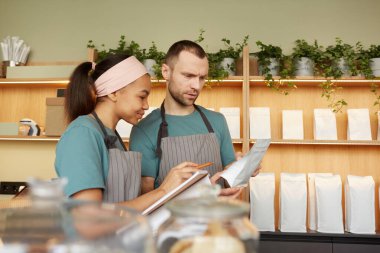 Waist up portrait of two young waiters wearing aprons while doing inventory in cafe or coffee shop, copy space