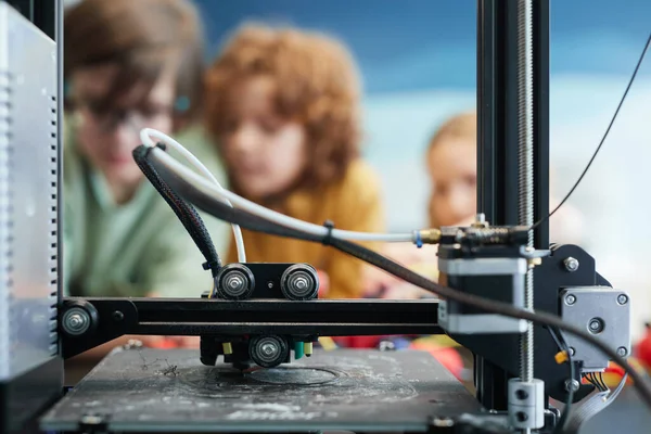 Close up of 3D printer making plastic models during engineering and robotics class at school with children in background, copy space