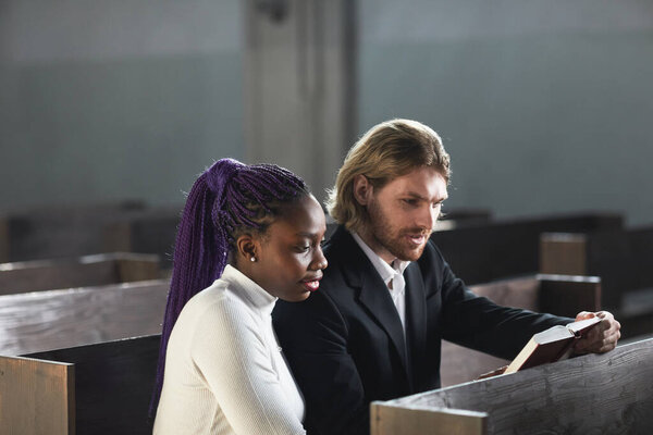 Multiethnic young couple sitting on the bench together and reading the Bible together in the church