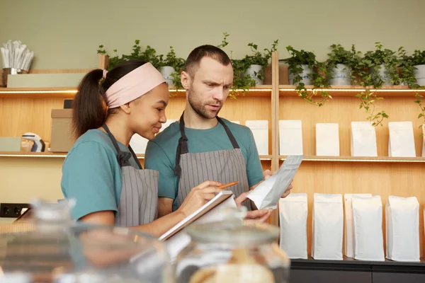 Waist up portrait of two young waiters wearing aprons while doing inventory in cafe or coffee shop, copy space