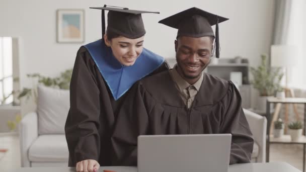卒業生のガウンを身に着けている若い混合人種の女性とアフリカの男性の中程度のショットとラップトップを使用して自宅でオンライン卒業式を持っている帽子 — ストック動画