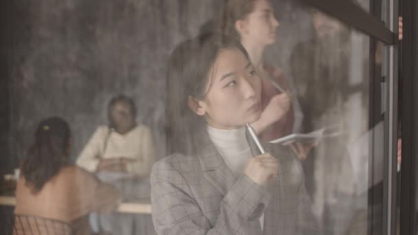 Pohled do truhly přes skleněnou stěnu zamyšlené asijské podnikatelky držící pero v ruce, dívající se na poznámky, přemýšlení a psaní v zaneprázdněné moderní kanceláři