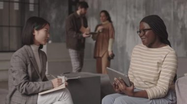 Modern ofiste oturmuş gülümseyen ve sohbet eden iki Asyalı ve Afrikalı kadın meslektaşın orta yavaş filmi. Arka planda duran iki bulanık iş arkadaşı.